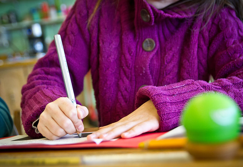 Élève assise à un pupitre en bois. Elle tient un crayon dans sa main et est en train d'écrire dans son cahier.